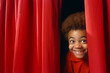 © Pics_With_Care - Young African American boy excited, peeking through red stage curtains, awaiting his moment in the school theatre play. Copy Space.