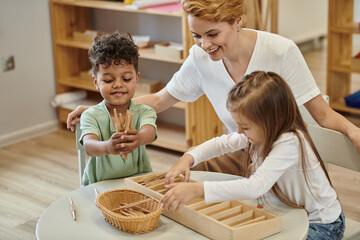 positive multiethnic kids playing with wooden didactic materials near teacher in montessori school