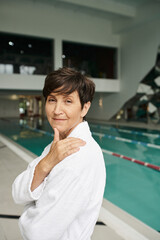 Wall Mural - happy middle aged woman with short hair in white robe, spa center, indoors swimming pool, smile