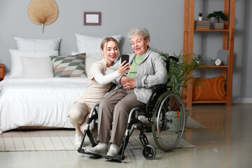 Wall Mural - Young caregiver with senior woman in wheelchair using mobile phone at home