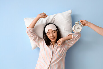 Wall Mural - Beautiful Asian woman in pajamas with pillow and ringing alarm clock on blue background