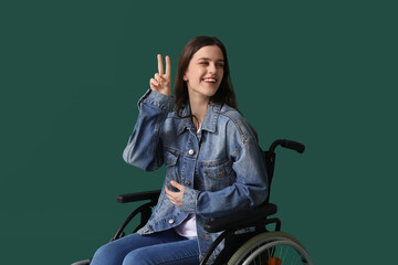 Wall Mural - Young woman in wheelchair showing victory gesture on green background