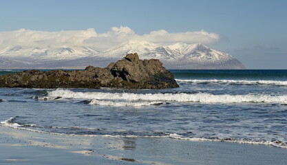 Wall Mural - Russia. Far East, Kuril Islands. View of the cold waves of the Pacific Ocean against the background of snow-capped volcanoes surrounded by basalt rocks.