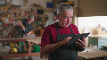 Senior worker wearing apron standing inside carpenter workshop holding tablet and browsing orders, job occupation concept using modern technology