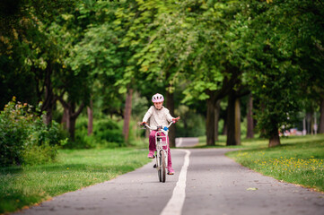 Little schooler girl riding bike in parks. Summer time, wearing helmet. From the back.