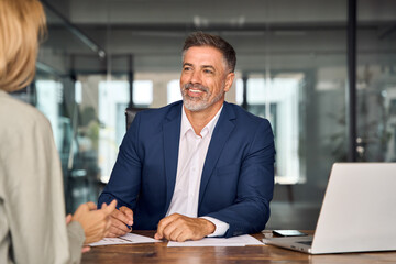 Wall Mural - Lawyer or real estate agent make consultation for client. Mature Latin business man and European business woman discussing project on laptop sitting at table in office. Business people work together.