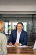 © Stock 4 You - Lawyer or real estate agent make consultation for client. Mature Latin business man and European business woman discussing project on laptop sitting at table in office. People work together, vertical.