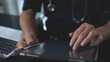 © tippapatt - Male doctor finger touching on on digital tablet screen and working laptop computer on table at doctor's office, close up, health technology, healthcare and medicine concept.