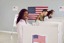 Side View Portrait Of A Happy Smiling American Voter Woman Making Thumb Up Gesture Looking Cheerful At Camera Voting And Putting Her Ballot In Bin On Election Day Standing At Vote Center.