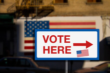 Vote Here Sign. With American Flag And Blurred Background. Concept Image.