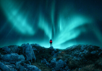 Wall Mural - Aurora borealis and young woman on mountain peak at night. Northern lights, stones and silhouette of alone girl on mountain trail. Landscape with polar lights. Starry sky with bright aurora. Travel
