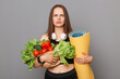 Sad unhappy upset young woman holding vegetables and yoga mat isolated on gray background looking at camera with frowning face expressing negative emotions.
