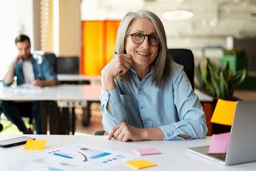 Portrait of smiling confident senior businesswoman using sticky notes looking at camera sitting in modern office. Gray haired manager wearing stylish eyeglasses  at workplace. Scrum, agile concept 