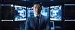 © Justlight - An Asian man stands proudly in front of a computer a wide spectrum of monitors behind him. He wears a modern grey suit with a navy blue tie looking intently and thoughtfully at the