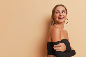 Wall Mural - Young woman with dreads wearing black top stands head to shoulder pose hugging herself on light brown backdrop, happiness concept, copy space