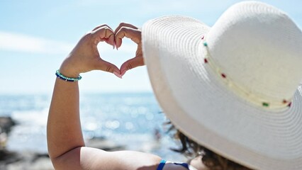 Canvas Print - Young beautiful latin woman tourist wearing summer hat doing heart symbol with hands at beach