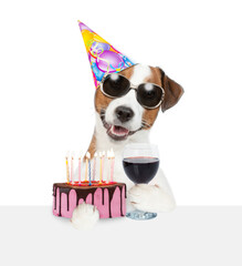Poster - Jack russell terrier puppy wearing a party hat holds birthday cake with lot of candle and glass of red wine above empty white board. isolated on white background