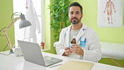 Canvas Print - Young hispanic man doctor using laptop holding medication bottle at clinic