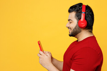 Wall Mural - Side profile view young happy Indian man he wears red t-shirt casual clothes listen to music in headphones use mobile cell phone isolated on plain yellow orange background studio. Lifestyle concept.