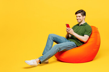 Wall Mural - Full body side view young happy man he wearing green t-shirt casual clothes sit in bag chair hold in hand use mobile cell phone isolated on plain yellow background studio portrait. Lifestyle concept.