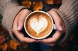 Nahaufnahme, Herbstliche Gemütlichkeit, Hände einer Frau mit Strickpullover halten eine Tasse Kaffee mit herzförmigen Milchschaum vor Herbsthintergrund mit Laub