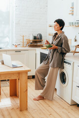 Wall Mural - A woman standing in the home kitchen eats a salad and looks at the laptop screen.