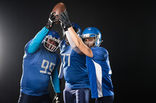 Three American Football Players Raising Their Hands Up Holding The Ball On A Black Background. 