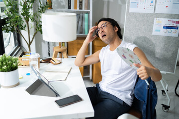 Asian Businessman blowing at herself with electric fan while sitting at workplace with documents and laptop.