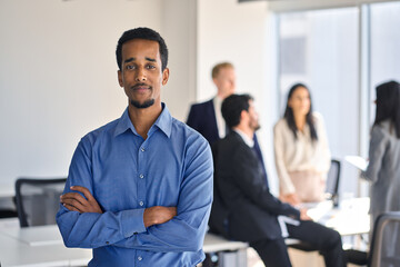 Poster - Confident young African American business man worker standing in office looking at camera. Smiling businessman employee corporate leader executive in meeting room with team, portrait.