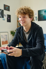 Wall Mural - Happy teenage guy in black hoodie and pants sitting on bed in front of computer and pressing console buttons while playing video game