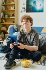 Sticker - Cheerful teenage boy sitting on the floor in front of glass bowl with potato chips while pressing buttons on controllers during video game