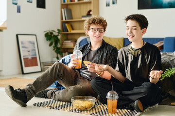 Sticker - Cute smiling teenage girl sitting in yoga pose on the floor next to guy eating potato chips, drinking soda or juice and looking at her