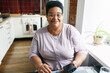© shurkin_son - Horizontal image of smiling plus size grandma of african ethnicity sitting at kitchen table with smartphone in hands next to window, having rest after household chores, looking at camera