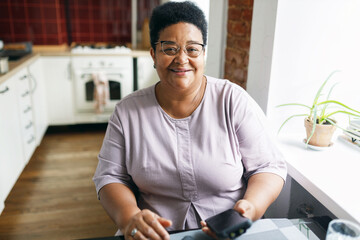 Horizontal image of smiling plus size grandma of african ethnicity sitting at kitchen table with smartphone in hands next to window, having rest after household chores, looking at camera