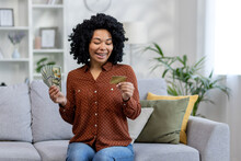 Young Happy African American Woman Holding Looking At Credit Card And Holding Cash Money And Bills In Hands, Sitting On Couch At Home