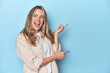 © Asier - Blonde young caucasian woman in blue studio pointing with forefingers to a copy space, expressing excitement and desire.