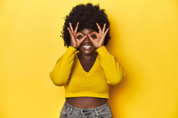 Wall Mural - African-American woman with afro, studio yellow background showing okay sign over eyes