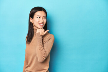 Wall Mural - Young Asian woman in brown turtleneck, points with thumb finger away, laughing and carefree.