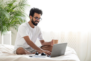 Wall Mural - Young Indian Man Using Laptop And Drinking Coffee While Sitting In Bed