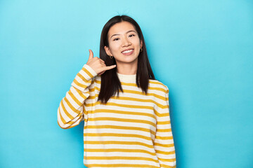 Wall Mural - Asian woman in striped yellow sweater, showing a mobile phone call gesture with fingers.