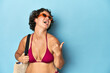 © Asier - Young woman in bikini with beach bag points with thumb finger away, laughing and carefree.