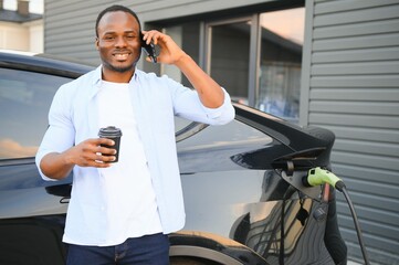 African man standing near electric car with charging cable in plug. Eco friendly vehicle charging on station. LIfestyles concept