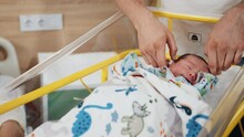 Baby In Hospital Crib, Father Putting His Hand Oh Her Head. A Newborn And His Parent At The Maternity Ward. Infant Baby Boy At Hospital Crib. Unrecognizable Father Make With Hands Protect Gesture