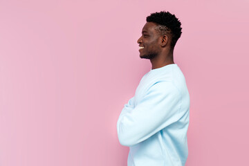 Sticker - african man in blue sweater smiles and stands with his arms crossed on pink isolated background, side view