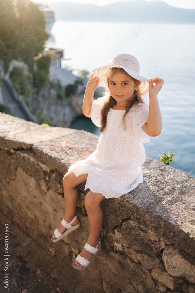 Cute little girl in beautiful white dress and hat sitting on bordure on ...