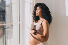 Charming Lovely Pregnant Female In Crop Top Patting Her Belly Standing In Front Of Window Under Sun Rays, Drinking Coffee Or Tea In Morning, Talking Too Her Baby, Imagining Their Future Life Together