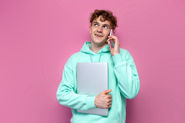 Wall Mural - young curly milennial guy in mint hoodie holds laptop and talks on the phone on pink isolated background