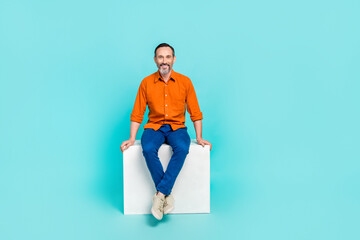 Full length photo of charming funny man dressed orange shirt sitting white cube isolated turquoise color background