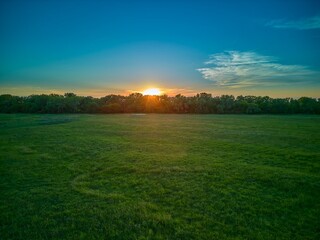 Wall Mural - Drone aerial view  of sun setting behind trees with large open field.