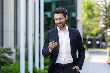 A young man businessman walks down the street in a suit and reads messages, news on the phone. He looks at the screen with a smile, calls, chats